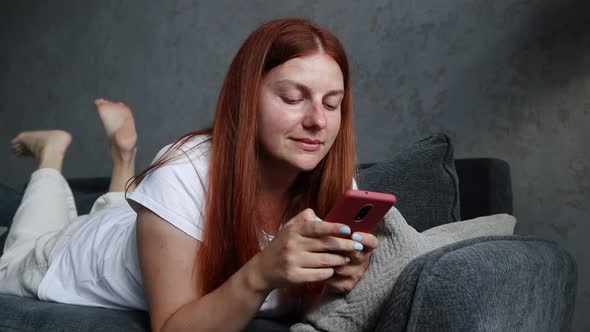 Beautiful Young 30s Redhaired Smiling Girl Lying on the Couch and Uses a Smartphone at Home Living