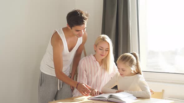 Young Parents Helping Cute School Child Girl Doing Homework Sitting at Kitchen Table