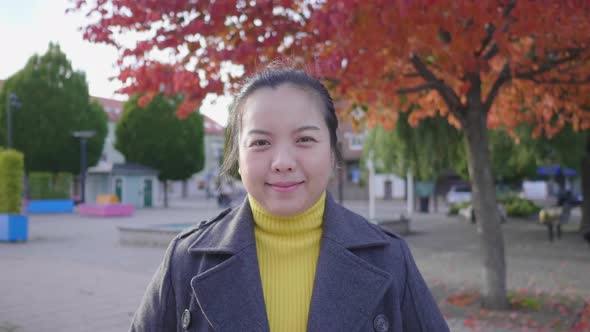 Front view of happy Asian woman standing in the park with red and green trees alt