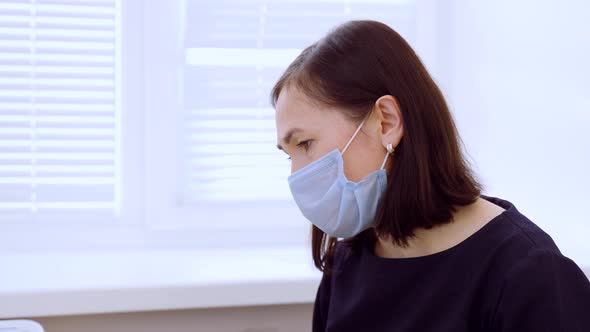 A Woman in a Medical Protective Mask Sits at a Table Against the Background of a Window alt