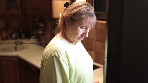 Hungry middle aged woman opens a refrigerator at night and eating hamburger with sausage. alt