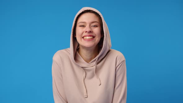 Young Happy Woman in Beige Hoodie Laughing Against Blue Background