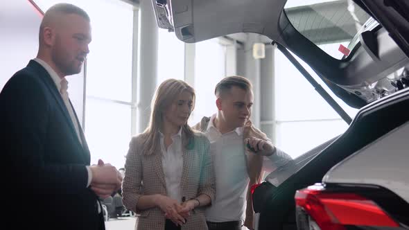 Professional Car Salesman is Telling Interested Buyers Couple About Luxurious Car with Trunk Open in alt