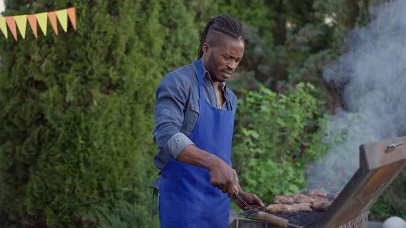 Middle Shot of African American Man in Blue Apron Checking Bbq Fire Cooking Burger Patties in Summer alt