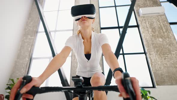 Young Female in VR Glasses is Pedaling Indoor Cycle Trainer alt
