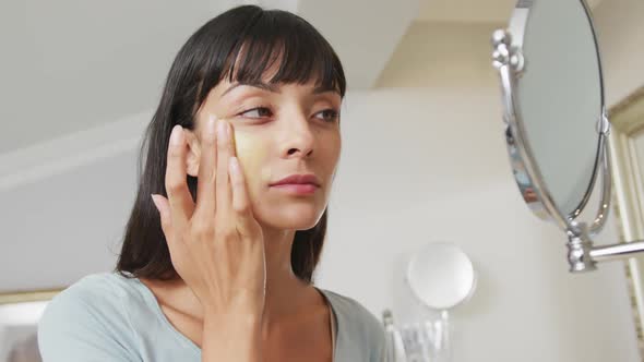 Caucasian woman looking in mirror and using cream on face in bathroom alt