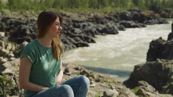 Woman Sits on Shady Bank of Narrow Mountain River in Summer alt