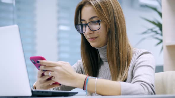 Female Office Worker Uses Phone While Working on Laptop Plays Mobile Game Break alt