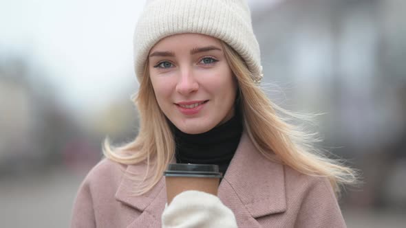 Young girl in a coat and hat standing on street and holding cup of hot drink. alt