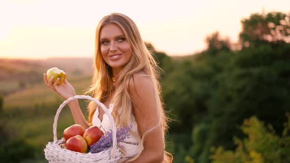 Beautiful sexy blonde girl in white dress posing in a field at sunset with a basket of fruit alt