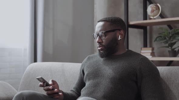 Handsome Young African American Guy Sitting on Couch in Living Room Wearing Wireless Bluetooth alt