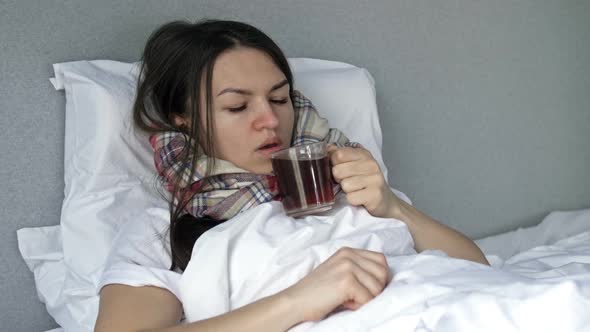 Sick Woman Lies in Bed Wrapped in a Scarf and Drinking Herbal Tea