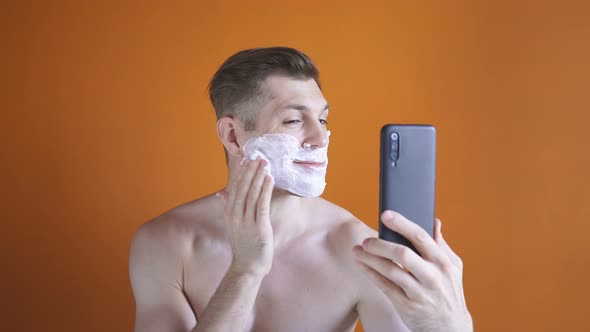 Young Man with White Shaving Foam on His Face Looking at His Smartphone, Isolated Background, Bare alt