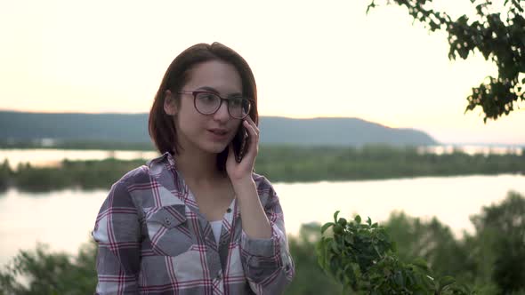 A Young Woman Stands on a Hill Against the Background of the River and Mountains and Speaks on the alt