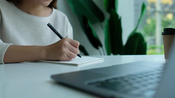 Closeup of a woman writing on a blank notebook with laptop computer on the table