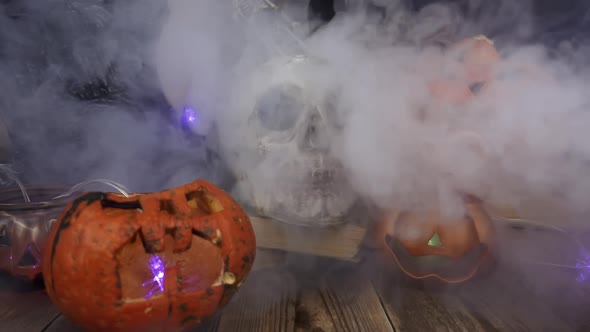Smoke Is Slowly Blowing at Pumpkins and Skull Standing at Wooden Table alt