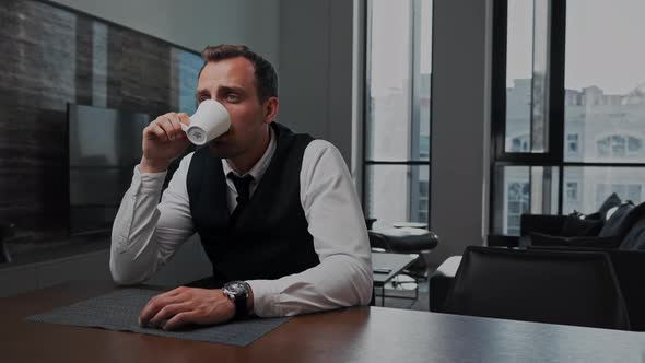 Business Man Sitting at the Table in the Office Drinks Coffee From a White Cup alt