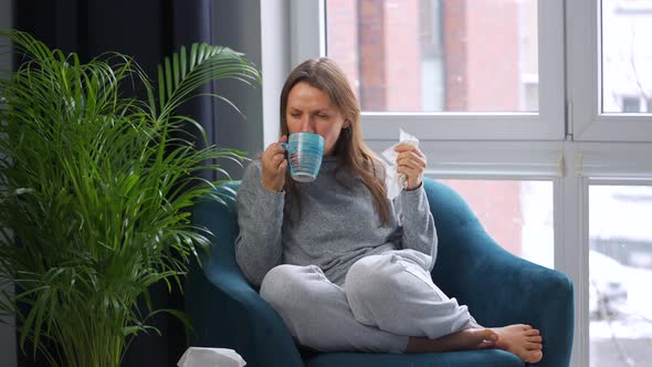 Unhealthy Woman Sits in a Chair Drinks Hot Tea or a Cold Medicine and Sneezes or Blows Her Nose Into alt