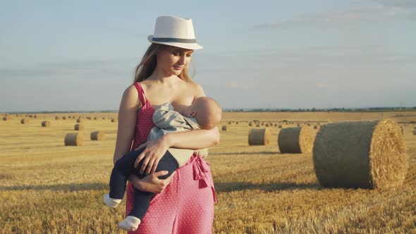 Happy Young Mother in Hat Feeds Baby Son with Breast in Haystack Field in Summer alt