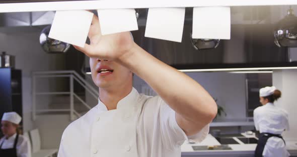 Caucasian male chef working in a restaurant kitchen checking orders, with colleagues working alt
