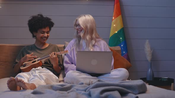 Young Lgbt Couple Sit in Bed Working on a Laptop and Playing on Ukulele