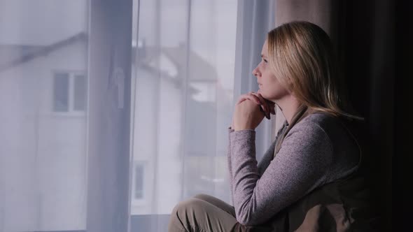 A Sad Woman Sits on the Window Sill, Looking Out the Window at the House alt