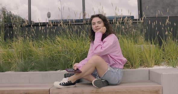 Portrait of joyful woman wearing pink shirt using a mobile phone, sit at the bus stop. alt