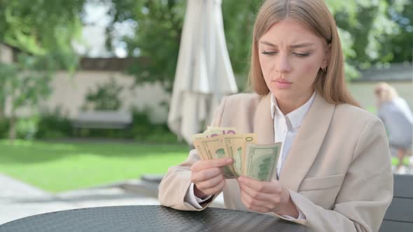 Upset Young Businesswoman Counting Dollars in Outdoor Cafe alt