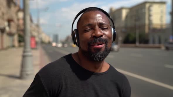 Happy Afro-american Man Is Walking on City Streets, Listening To Music By Headphones and Dancing alt