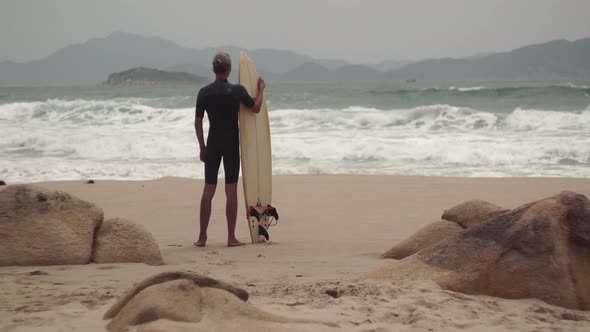 Man with Surfing Board Looks at Sea Waves Standing on Beach
