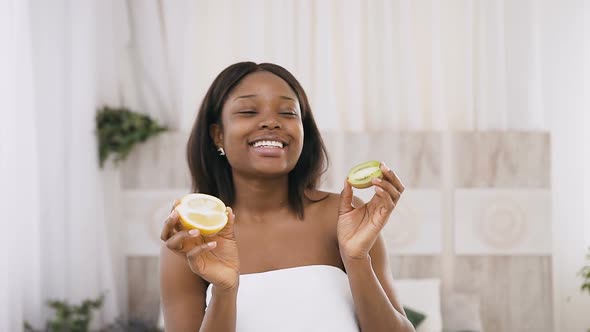 Funny Amusing African American Young Woman Holding Kiwi and Orange Halves in Hands Over Light alt