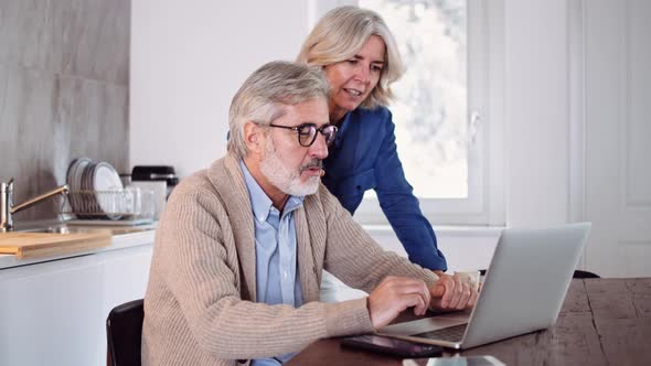 Slow motion shot of mature couple using laptop at table in the kitchen alt