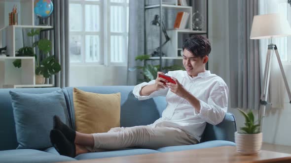 Asian Man Playing Game On Smartphone And Smiling While Lying On Sofa In The Living Room alt