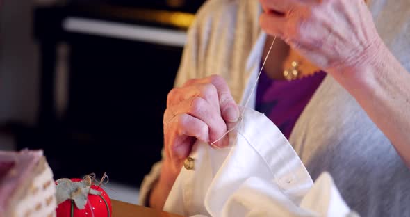The wrinkled hands of an aging elderly woman sewing a button onto a white dress shirt with thread. alt