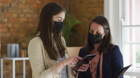 Caucasian female business owner and her coworker wearing face masks, using tablet and talking alt