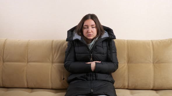 Frozen Woman in Warm Jacket Tries to Heat Palms on Sofa alt