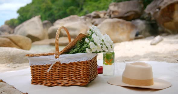 Picnic Basket with Flowers Straw Hat Bottle of Fresh Lemonade are Standing on Tablecloth on Beach alt