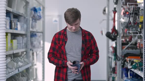 Portrait of Young Caucasian Man Looking in Empty Wallet Standing Between Shelves in Hardware Store alt
