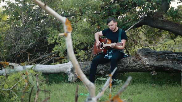 A Young Man in a Black Tshirt Sits on a Felled Tree and Slowly Plays an Acoustic Guitar