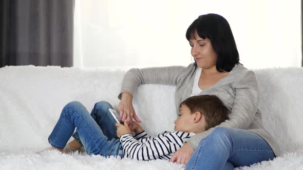Happy Family: Mother and Son Spend Time Playing with a Tablet. Woman and Child Sitting on the Sofa alt