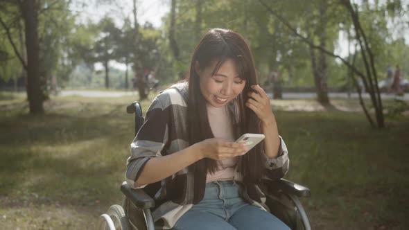 A Young Disabled Asian Girl with Long Dark Hair is Sitting in the Wheelchair in a Public Park alt