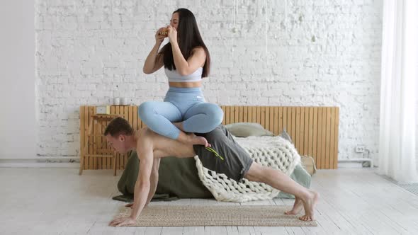 Man Is Doing Static Plank Exercise at Home with Woman Sitting Above on His Back