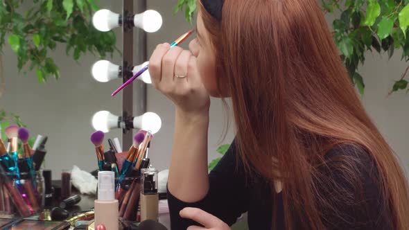 Young Woman Applying Makeup While Sitting at Her Vanity Table with Lots of Cosmetics alt