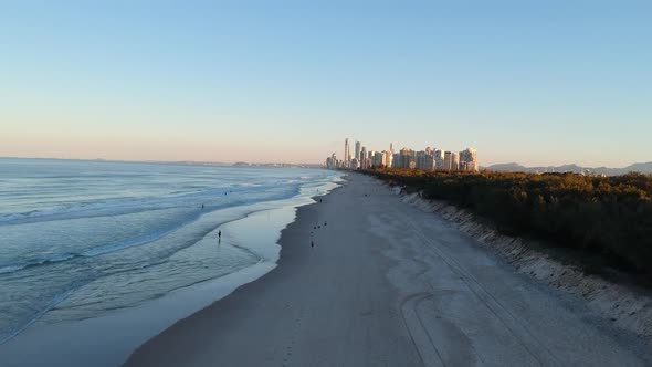 Sun setting on a beach while people walk with the city skyline glistening in the distance alt