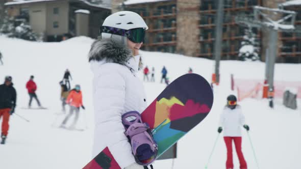 Woman with Snowboard Walking By Snowy Mountain with Skiers Riding Down Slope alt