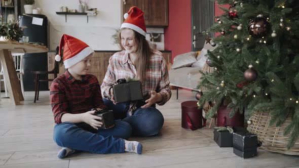 Happy Caucasian Mom and Son Sit Near Christmas Tree and Exchange Gifts in Boxes alt