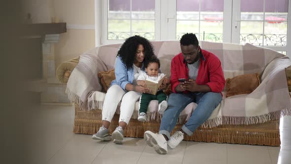 Wide Shot Portrait Loving African American Mother Reading Book to Cute Son As Father Messaging alt