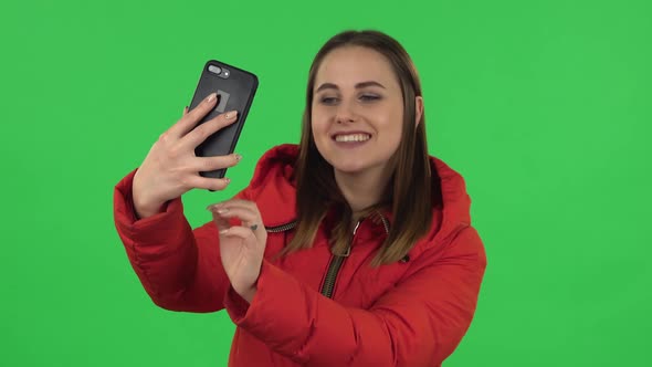 Portrait of Lovely Girl in a Red Down Jacket Is Making Selfie on Mobile Phone Then Looking Photos alt