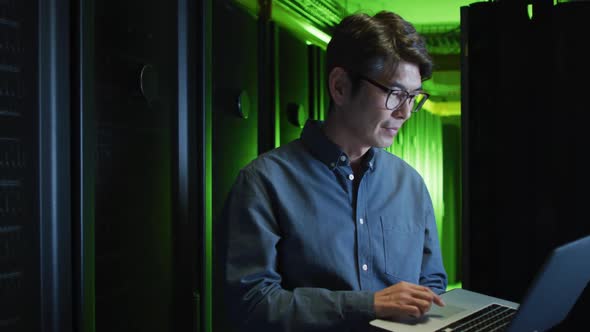 Smiling asian male it technician using laptop checking computer server alt