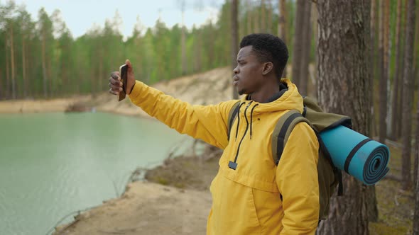 A Black Man with a Backpack Uses a Mobile Phone While Hiking Through the Forest in the Mountains alt